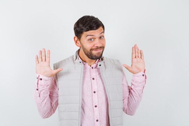 Knappe jonge man toont overgave gebaar in shirt, vest en ziet er vrolijk uit, vooraanzicht.