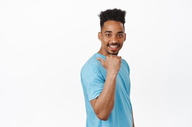 Knappe jonge mannelijke modelmodel, Afro-Amerikaanse man in t-shirt wijzende vinger naar links, achter zijn schouder, glimlachend en gelukkig kijken, uitnodigen voor evenement op wit