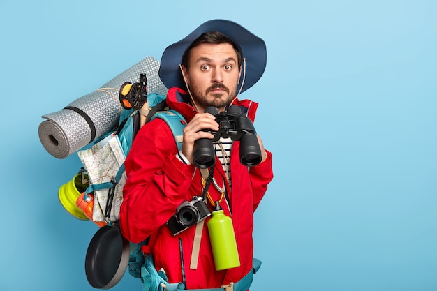 Gratis foto knappe jonge mannelijke toerist houdt een verrekijker, wandelingen in het bos, draagt vrijetijdskleding, draagt noodzakelijke dingen om te reizen, brengt graag vakantie door in actieve kleding