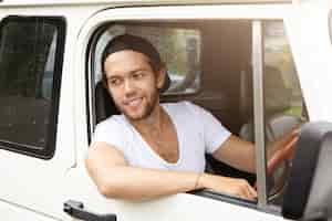 Gratis foto knappe jonge mens die honkbal glb dragen die wit suv-voertuig achteruit drijven, zijn elleboog uit open venster steken, het glimlachen