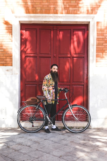 Gratis foto knappe jonge mens met fiets die zich voor houten rode deurmuur bevindt