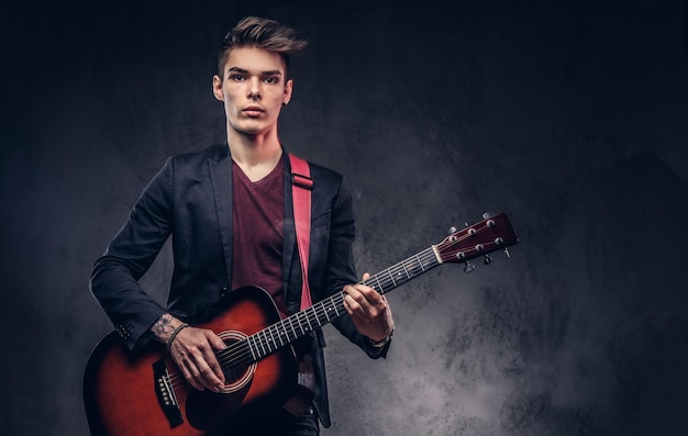 Knappe jonge muzikant met stijlvol haar in elegante kleding met een gitaar in zijn handen spelen en poseren op een donkere achtergrond.