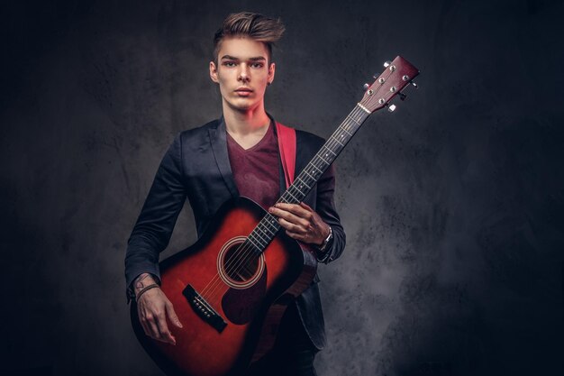 Knappe jonge muzikant met stijlvol haar in elegante kleding met een gitaar in zijn handen spelen en poseren op een donkere achtergrond.