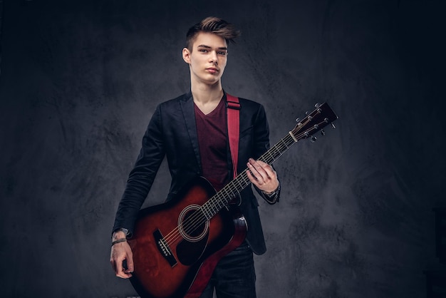 Knappe jonge muzikant met stijlvol haar in elegante kleding poseren met een gitaar in zijn handen op een donkere achtergrond.