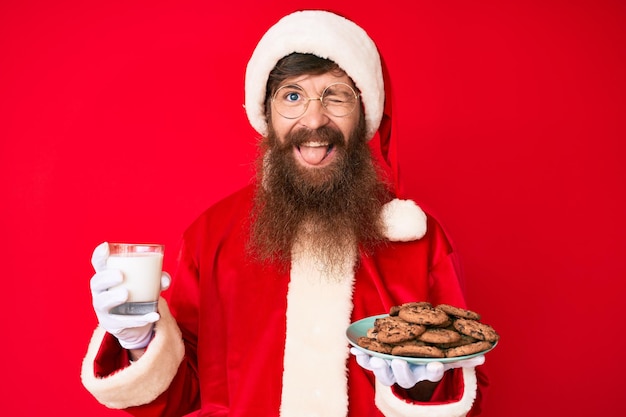 Knappe jonge roodharige man met lange baard in kerstmankostuum met koekjes en melk knipogend kijkend naar de camera met sexy uitdrukking, vrolijk en blij gezicht.