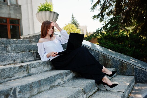 Knappe jonge vrouw in witte blouse, wijde zwarte broek en zwarte klassieke hoge hakken, zittend op de trap en werkend op haar laptop