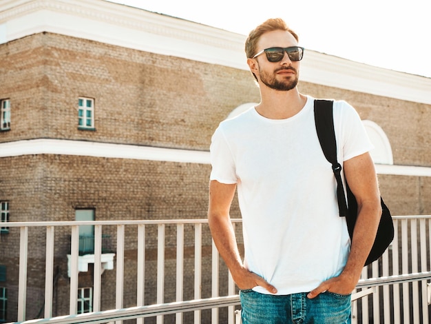 Knappe lachende hipster lamberseksueel modelstijlvolle man gekleed in witte tshirt en spijkerbroek mode man poseren op de straat achtergrond in de buurt van hek op het strand met schooltas in zonnebril