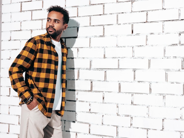 Knappe lachende hipster model Sexy ongeschoren man gekleed in gele zomer shirt en jeans kleding mode man poseren in de buurt van witte bakstenen muur in studio op zonnige dag