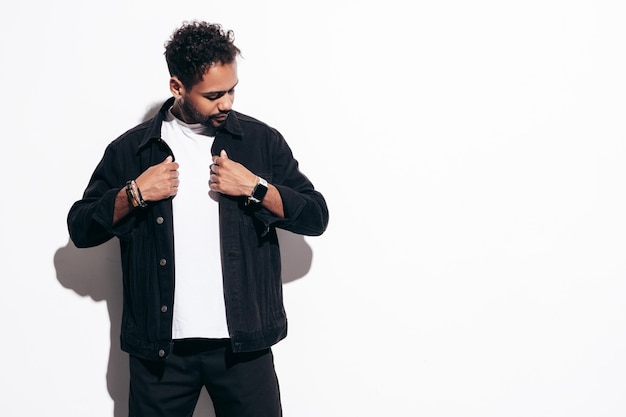 Knappe lachende hipster model Sexy ongeschoren man gekleed in zwarte zomer shirt en jeans kleding mode man poseren in de buurt van witte muur in studio op zonnige dag