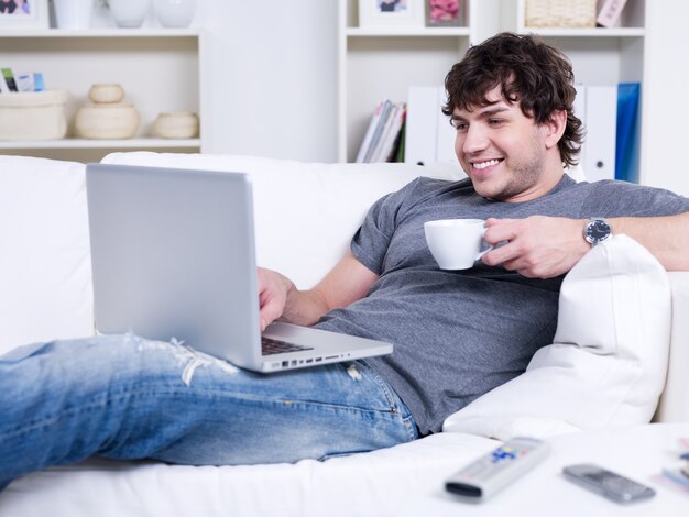 Knappe lachende jonge man met kopje koffie met laptop thuis