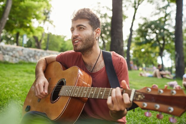 Knappe lachende man gitaarspelen in park, zittend op het gras, zorgeloos weekend hebben