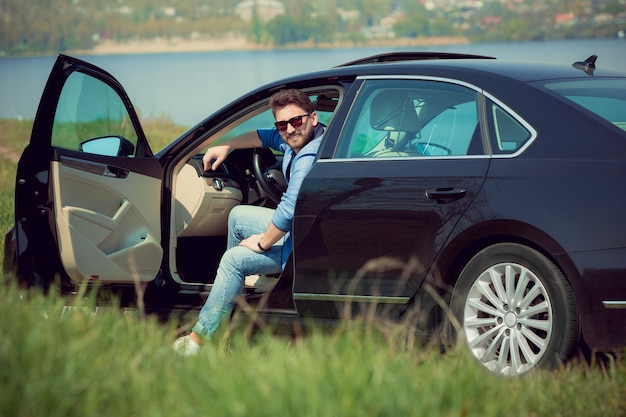 Knappe lachende man in spijkerbroek, jas en zonnebril zit in zijn auto met geopende deuren aan de kant van de rivier.