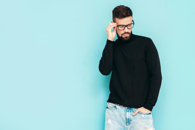 Knappe lachende model Sexy stijlvolle man gekleed in zwarte coltrui en jeans Fashion hipster man poseren in de buurt van blauwe muur in studio geïsoleerd In bril of bril