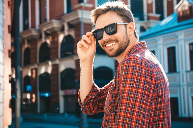 Knappe lachende stijlvolle hipster lamberseksueel model. Moderne man gekleed in rood geruit overhemd. Mode man poseren in de buurt van wolkenkrabber op de straat achtergrond in zonnebril. Buiten bij zonsondergang