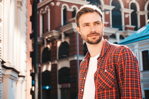 Knappe lachende stijlvolle hipster lamberseksueel model. Moderne man gekleed in rood geruit overhemd. Mode man poseren in de buurt van wolkenkrabber op de straat achtergrond in zonnebril. Buiten bij zonsondergang