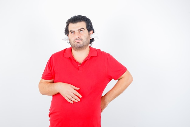 Knappe man die hand achter de taille houdt, aan iets denkt, met servetten in de oren in rood t-shirt staat en peinzend kijkt, vooraanzicht.