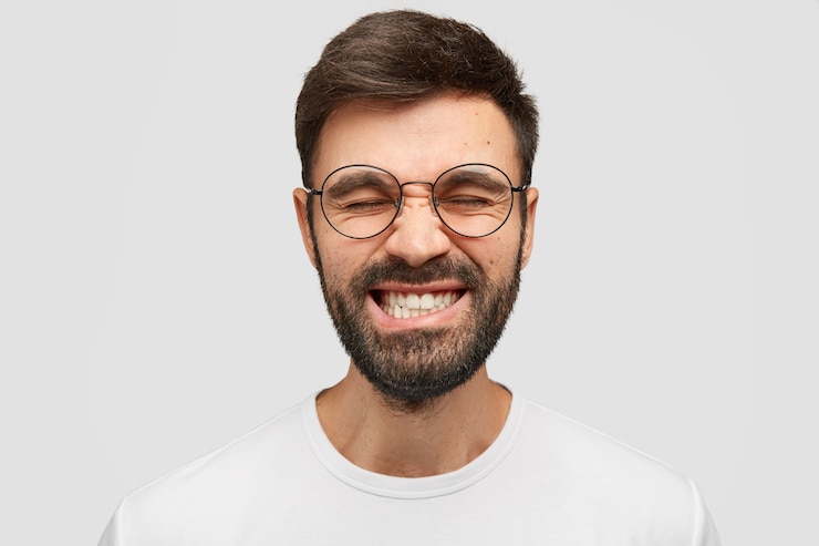 Gratis Foto knappe man fronst zijn wenkbrauwen en klaart zijn tanden, houdt de ogen gesloten, probeert zich ergens op te concentreren, draagt casual t-shirt