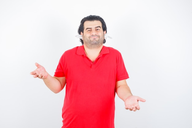 Knappe man in rood t-shirt met hulpeloos gebaar, staande met servetten in de oren en verbaasd, vooraanzicht op zoek.