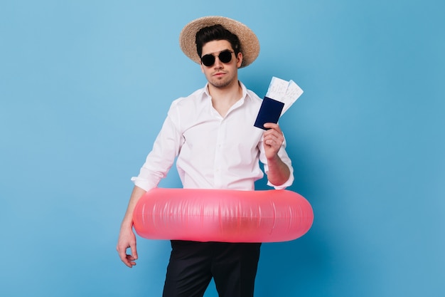 Knappe man in strooien hoed toont paspoort en vliegtickets. Portret van een man in zakelijke kleding met opblaasbare cirkel.