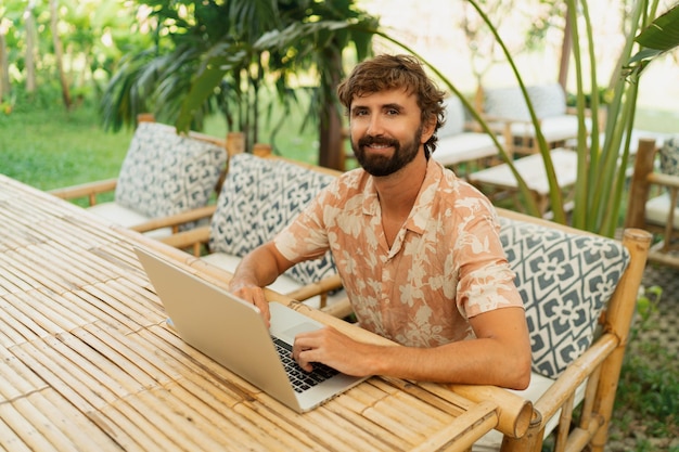 Knappe man met baard met behulp van laptop en mobiele telefoon zittend in terras met tropisch interieur