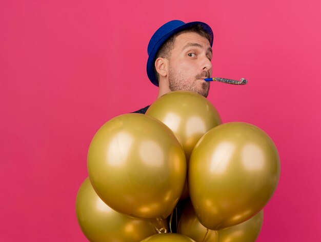 Knappe man met blauwe feestmuts houdt helium ballonnen blazen fluitje geïsoleerd op roze muur met kopie ruimte