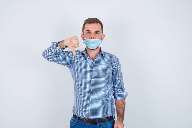 Knappe man met duim omlaag in shirt, spijkerbroek, masker en op zoek naar ontevreden, vooraanzicht.