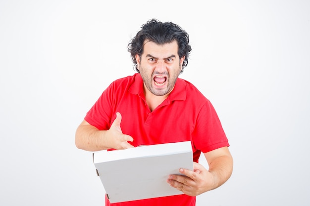 Knappe man met kartonnen doos, hand naar toe strekken met een boze manier in rood t-shirt en op zoek boos, vooraanzicht.