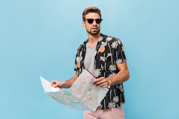 Knappe man met zwarte zonnebril in koel zomershirt en licht shirt wegkijkend en kaart vasthoudend op geïsoleerde achtergrond