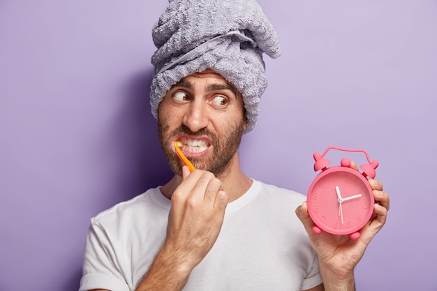 Knappe man poetst tanden, witt met tandpasta, houdt wekker in de hand, werd laat in de ochtend wakker, heeft een handdoek om het hoofd gewikkeld, draagt een casual wit t-shirt, geïsoleerd op een paarse muur. Ochtend routine