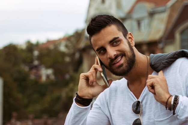 Knappe man praten buitenshuis aan de telefoon. Met leren jas, zonnebril, een man met baard. Instagram effect