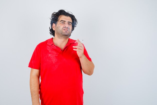 Knappe man roken sigaret in rood t-shirt en op zoek naar serieus. vooraanzicht.