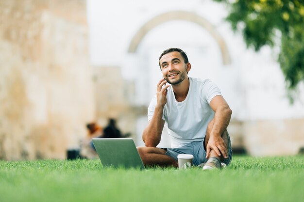 Knappe man zittend op het gras in de stad met een laptop en praten aan de telefoon