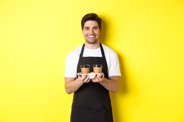 Knappe mannelijke barista die afhaalkoffie serveert en glimlacht, orde brengt, die zich in zwarte schort tegen gele achtergrond bevindt.