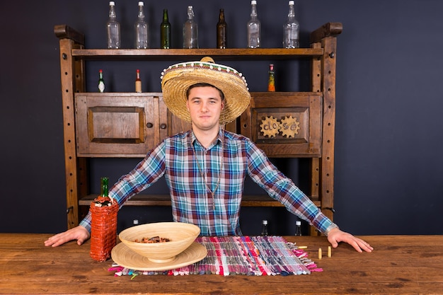 Knappe mannelijke barman in een sombrero die aan de balie staat, een fles tequila en een bord met snacks erop in de mexicaanse pub