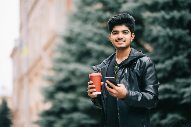 Knappe mensenstudent met oogglazen die met vrienden op mobiele telefoon spreken die van stedelijke koffie genieten terwijl het lopen op straat