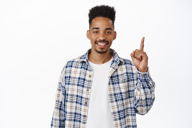 knappe moderne afro-amerikaanse jongen 20s jaar, wijzende vinger omhoog en glimlachend blij met witte tanden, reclame tonen, studio