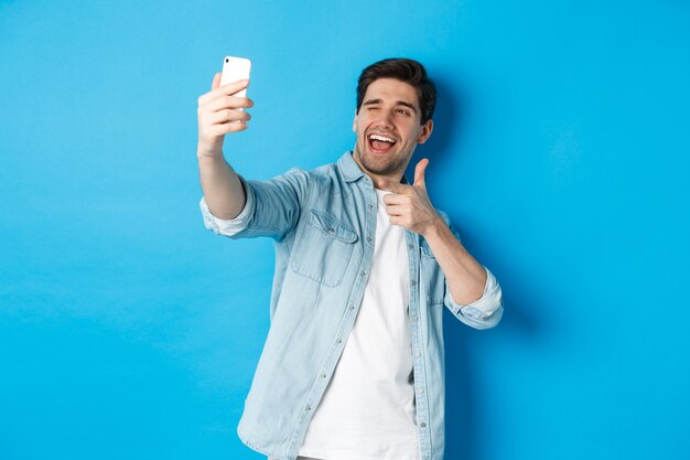 Knappe moderne kerel die selfie op smartphone neemt en vingerkanon richt op mobiele camera, brutaal knipoogt, staande tegen blauwe achtergrond