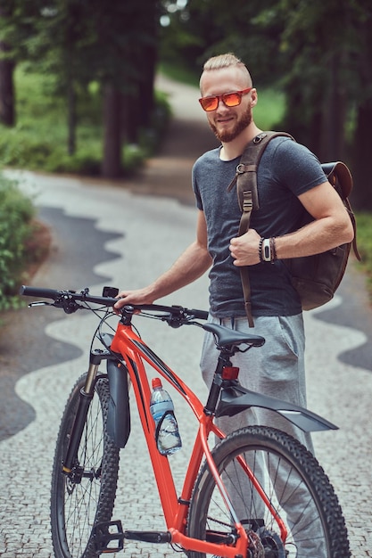 Knappe roodharige man met een stijlvol kapsel en baard gekleed in sportkleding en zonnebril loopt in het park met een fiets en rugzak.