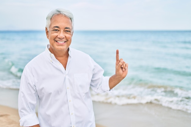 Gratis foto knappe spaanse man met grijs haar glimlachend gelukkig op het strand genietend van vakantie wijzend met de vinger naar de kopieerruimte