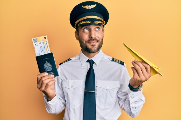 Knappe Spaanse piloot man met papieren vliegtuig en paspoort glimlachend naar de zijkant kijkend en denkend weg starend