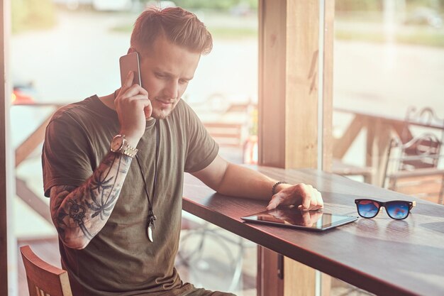 Knappe stijlvolle hipster zit aan een tafel in een café langs de weg, pratend op de smartphone.