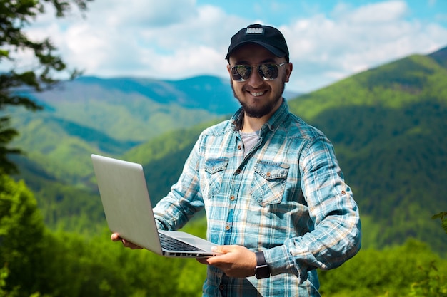 Knappe stijlvolle jongeman in zonnebril en met een laptop in handen kijkend naar de camera en glimlachend in de bergen