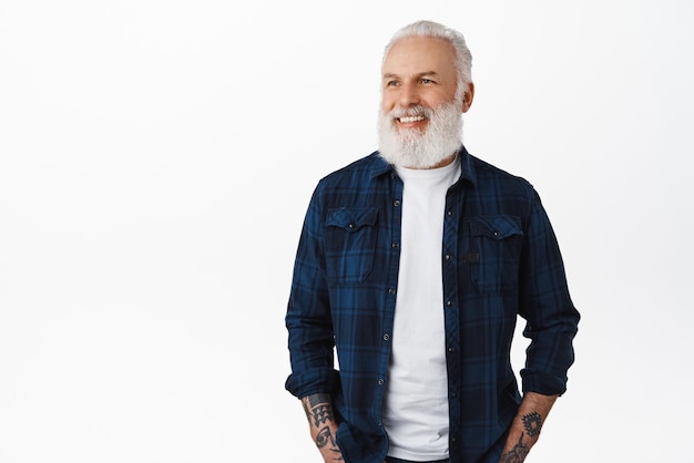 Knappe stijlvolle oude hipster man senior met witte baard en tatoeages opzij kijken naar advertentie met blij gezicht glimlachend gelukkig lezen promotionele tekst permanent over studio achtergrond