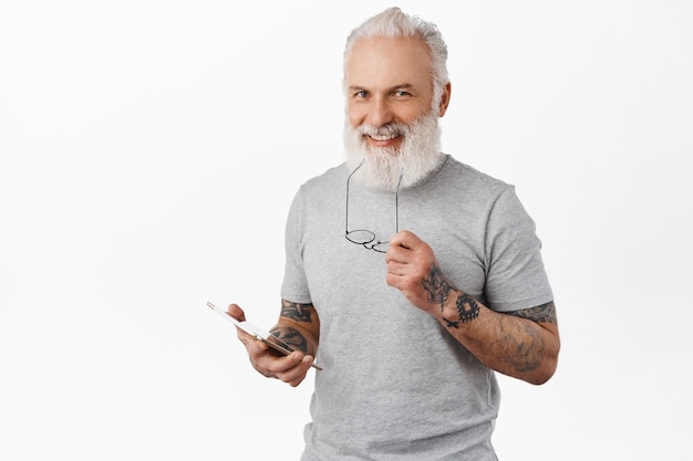 Knappe tevreden senior man met digitale tablet, bijten tempel van bril en tevreden glimlachen, online winkelen, berichten sturen op sociale media, staande over witte muur