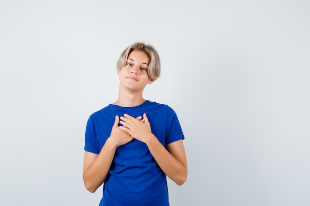 Knappe tienerjongen in blauw t-shirt die de handen op de borst houdt en er dankbaar uitziet, vooraanzicht.