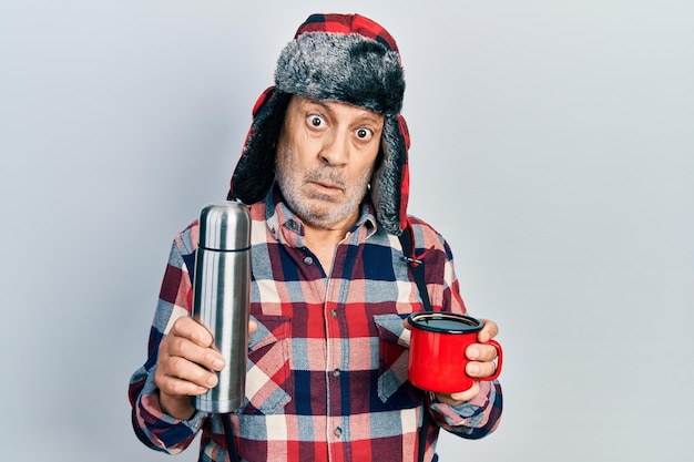 Gratis foto knappe volwassen klusjesman met wintermuts met oorflappen die hete koffie drinkt van thermo zonder idee en verwarde uitdrukking. twijfel concept.