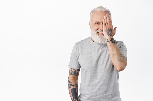 Knappe volwassen man met tatoeages, verbergt de helft van het gezicht, bedek het oog met de hand en lacht blij aan de voorkant, staande in grijs t-shirt tegen de witte muur