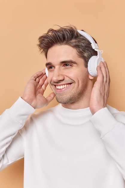 Knappe vrolijke Europese man glimlacht aangenaam houdt handen op draadloze stereo hoofdtelefoon heeft optimistische gezichtsuitdrukking geniet van het luisteren van muziek tijdens vrije tijd poses binnen. Hobbyconcept