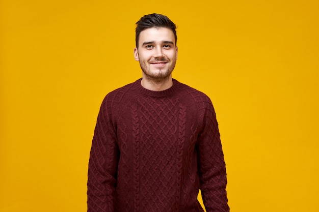 knappe vrolijke jonge man met stijlvol kapsel en kuiltjes glimlach poseren geïsoleerd tegen lege gele muur, gekleed in een gezellige kastanjebruine trui, met zelfverzekerde blik
