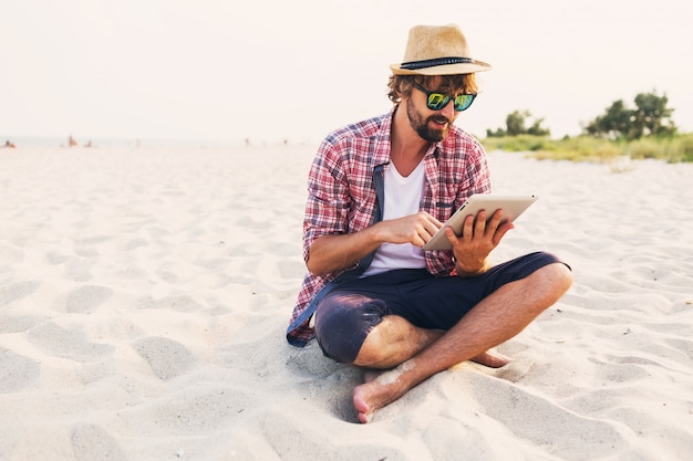 Knappe vrolijke man met baard in strohoed, geruit overhemd en stijlvolle zonnebril zittend op wit zand en tablet gebruiken